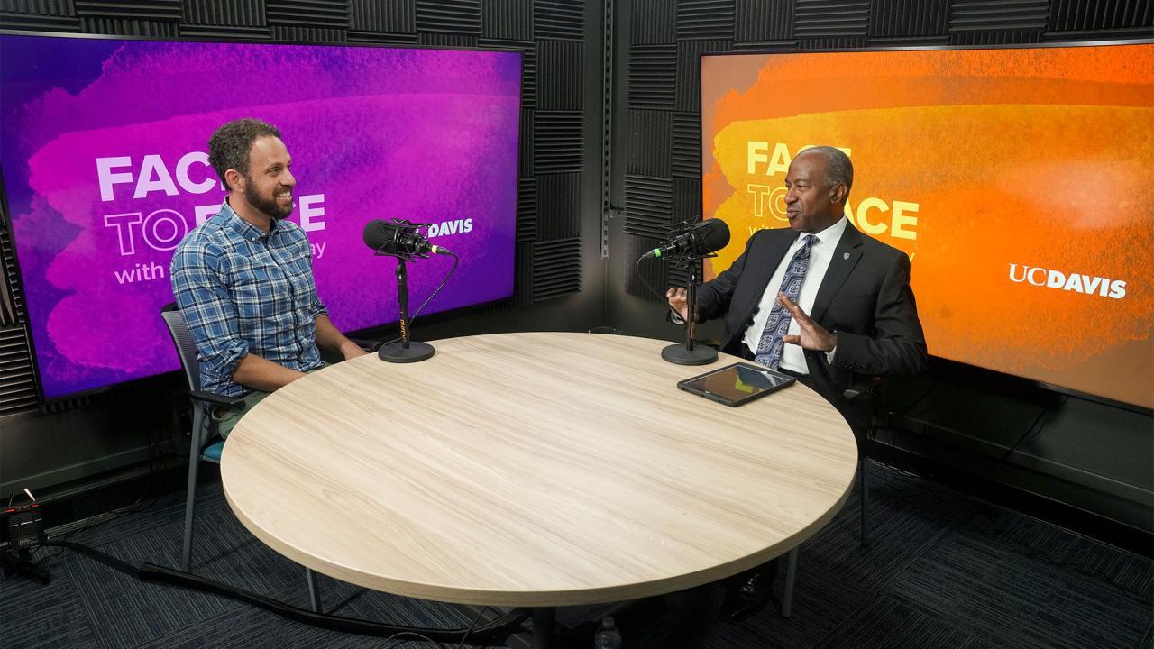 Two people sitting at a round wooden table with a purple screen behind one of them and an orange screen behind the other