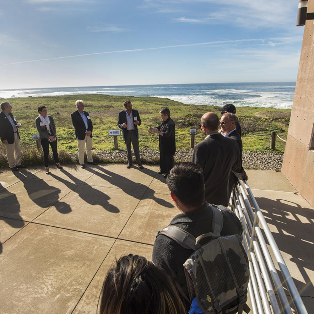 A group of people standing around watching one person present. They are outdoors, with a view of the coast and ocean in the background. Their shadows are stretching across the pavement because the sun is at their backs.
