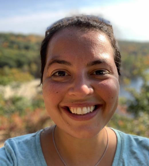 A close up portrait image of a person with dark, pulled back hair, smiling at the camera in front of a background of desert plants and blue skies with white, streaky clouds