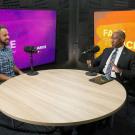 Two people sitting at a round wooden table with a purple screen behind one of them and an orange screen behind the other