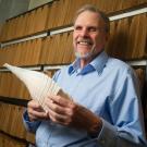 A person in a blue button up shirt standing in front of a row of brown wooden cabinets and holding a sheet of paper.
