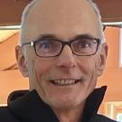 A person in a dark jacket and glasses standing in front of an orange background.